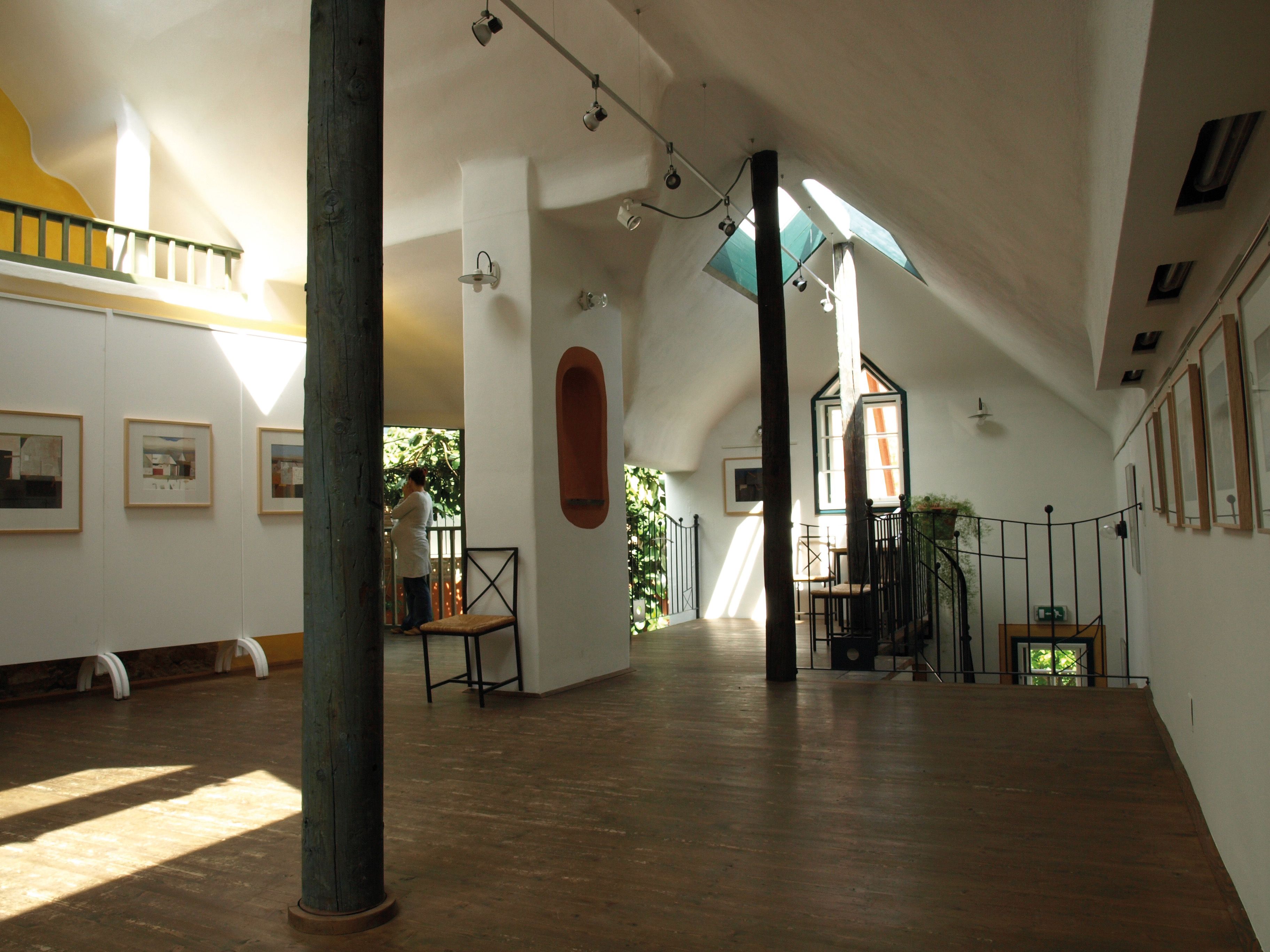 Innenansicht einer hellen Galerie mit Holzboden, Kunstwerken an den Wänden und einer Person am Fenster.