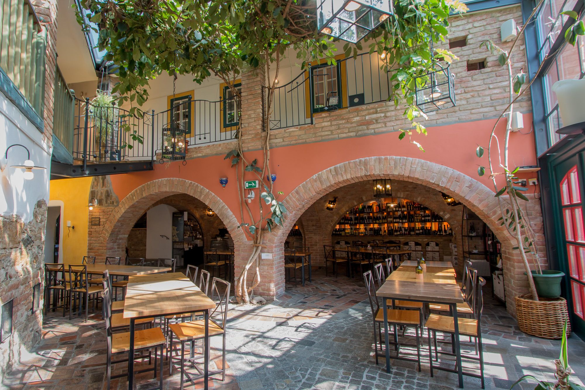 Innenhof eines Restaurants mit Tischen, Stühlen und Pflanzen, umgeben von Backsteinbögen und einem Balkon.