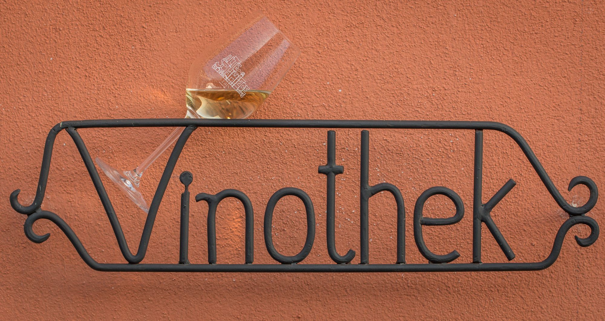 Metallschild mit der Aufschrift 'Vinothek' und einem Weinglas auf roter Wand.